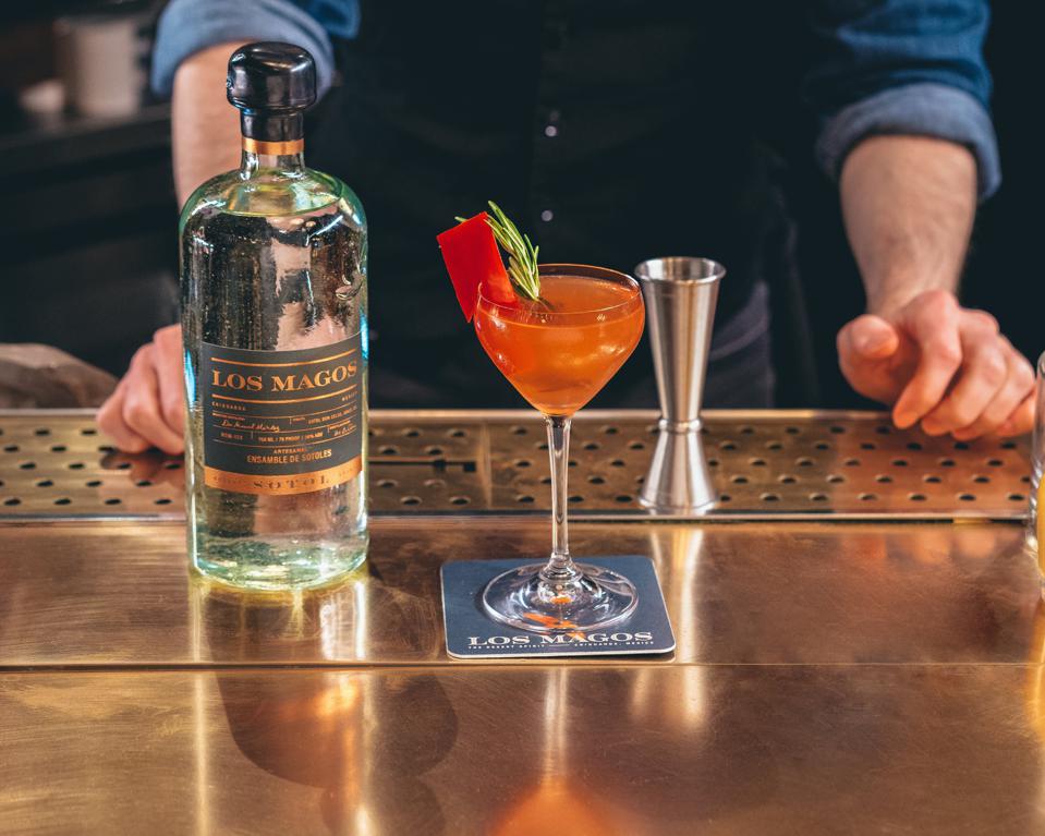 A bartender at a copper bar top with a bottle of Los Magos Sotol Blanco and a finished cocktail