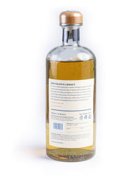A clear glass bottle of Los Magos Sotol 6 Year Reserva, Don Celso's Legacy sotol with a cork stopper, showing the back label and golden liquid inside, set against a plain white background.