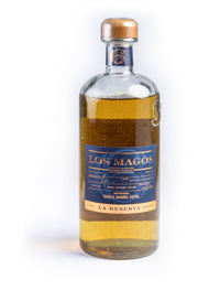 A bottle of Los Magos La Reserva Single Barrel Sotol, featuring a clear glass bottle with golden-yellow liquid inside, a blue label with gold lettering, and a light brown wooden cap, set against a plain white background.