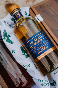 A bottle of Los Magos Reserva single barrel spirit with a blue and gold label, resting on a white branded paper towel inside a wooden box next to another bottle.