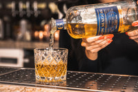 A bottle of Los Magos 6 Year Reserva Sotol being poured from a clear glass into a cut crystal tumbler on a bar counter, with a bartender's hands visible holding the bottle.